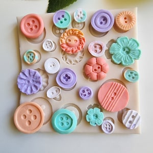 May include: A selection of colourful buttons in a silicone mould. The buttons are in shades of peach, lavender, mint green, and white. Some have floral designs, others classic shapes. The mould is on a white surface.