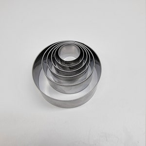 May include: A collection of six stainless steel, circular cookie cutters of different sizes, nested together. The cutters are arranged in a spiral, with the largest at the base and the smallest in the centre. The cutters are on a white background.