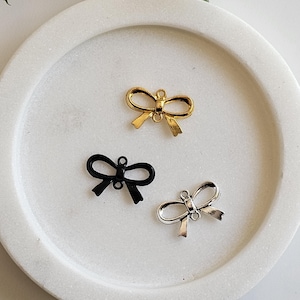 May include: Three metal bow charms in gold, black, and silver, displayed on a white, circular dish. The charms are small and detailed, with a loop at the top for attaching to jewellery or other crafts. Green foliage is in the background.