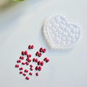 May include: A scattering of small, dark pink bow-shaped decorations on a white surface. A heart-shaped mould with a scalloped edge and bow-shaped impressions is also visible. The image has a clean, bright aesthetic, suitable for craft projects.