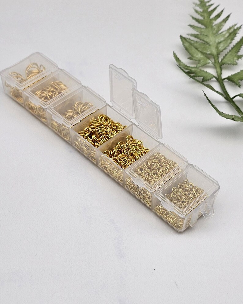 Op de afbeelding: Een doorzichtige plastic organizer gevuld met verschillende maten goudkleurige sprongringen. De organizer heeft meerdere compartimenten, elk met een ander type ring. Een takje groen blad is op de achtergrond.