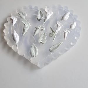 May include: A heart-shaped silicone mould with a scalloped edge, filled with white floral and leaf elements. The mould contains various flower buds and leaves, ideal for crafting projects. The design is simple and elegant.