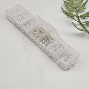 Op de afbeelding: Een doorzichtige plastic organizer gevuld met verschillende maten zilveren sprongringen. De sprongringen zijn netjes gerangschikt in afzonderlijke compartimenten. Een groen varenblad staat op de achtergrond.