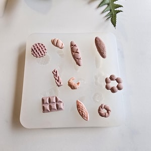 May include: A white silicone mould containing miniature bread shapes, including loaves, rolls, and a croissant. The bread shapes are a light brown colour. Green foliage is visible in the background.