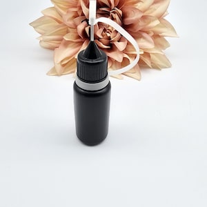 May include: A black plastic bottle with a white cap and a long, thin needle-like tip. The bottle has a white band around the middle. The bottle is sitting on a white surface.