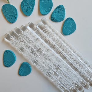 May include: Clear acrylic rolling pins with floral patterns and teal clay stamps with various designs. The tools are arranged on a white surface, ideal for crafting or baking.