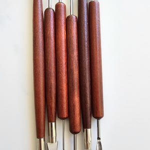 May include: A collection of seven pottery tools, each with a wooden handle and a stainless steel blade. The tools feature diverse shapes, such as loops, blades, and a needle. The handles are a deep, dark brown colour.
