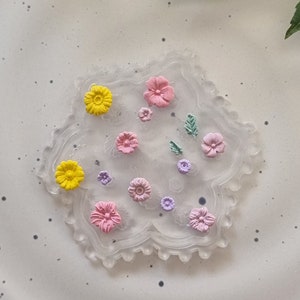 May include: A clear, decorative coaster with a scalloped edge, decorated with colourful, raised floral designs. The flowers are in shades of yellow, pink, and purple, with green leaf accents. A charming, handcrafted item.
