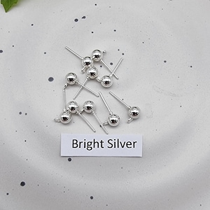 May include: A close-up of a group of silver ball head pins. The pins are scattered on a white surface with black speckles. The text "Bright Silver" is written on a white label.