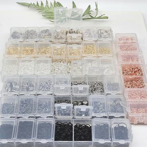 Op de afbeelding: Een verzameling kleine, doorzichtige plastic bakjes gevuld met verschillende maten en kleuren van jumpringen. De jumpringen zijn in tinten zilver, goud, zwart en koper. De bakjes zijn in een rasterpatroon gerangschikt.