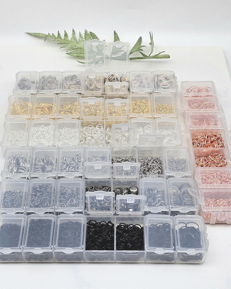 Op de afbeelding: Een verzameling kleine, doorzichtige plastic bakjes gevuld met verschillende maten en kleuren van jumpringen. De jumpringen zijn in tinten zilver, goud, zwart en koper. De bakjes zijn in een rasterpatroon gerangschikt.