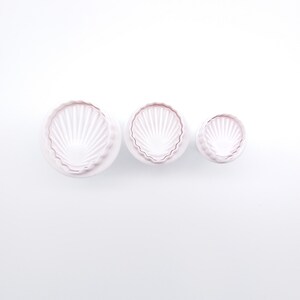 May include: Three light pink seashell-shaped cookie cutters of varying sizes. The cutters have a scalloped edge and a fan-like design inside. The cutters are arranged in a row against a white background, ideal for baking.