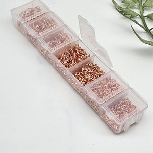 Op de afbeelding: Een doorzichtige plastic organizer gevuld met verschillende maten ros&eacute;gouden sprongringen. De organizer heeft meerdere compartimenten, elk met een andere hoeveelheid metalen ringen. Een takje groen is op de achtergrond.
