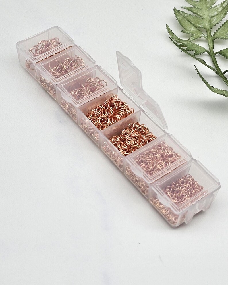 Op de afbeelding: Een doorzichtige plastic organizer gevuld met verschillende maten ros&eacute;gouden sprongringen. De organizer heeft meerdere compartimenten, elk met een andere hoeveelheid metalen ringen. Een takje groen is op de achtergrond.
