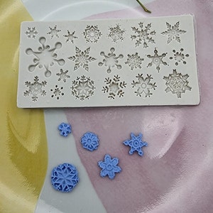 May include: A silicone mould with 20 snowflake designs. The mould is white and has a light grey background. There are five blue snowflake shapes on a light purple and yellow surface.