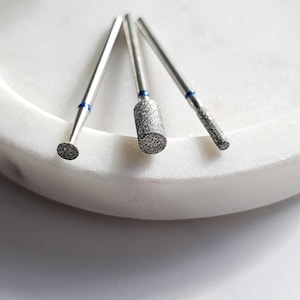 May include: Three silver-coloured metal nail drill bits with textured abrasive heads and thin shanks. Each bit features a small blue band near the base of the head. The bits include a small round tip, a medium cylindrical head, and a thin rod tip.