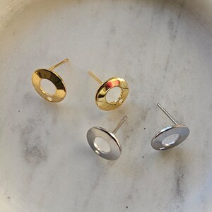 May include: Three pairs of earrings, two gold and one silver, with a circular design and a small hole in the centre. The earrings are displayed on a white marble surface.