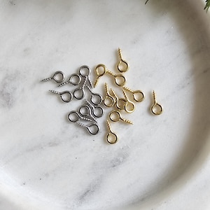 May include: A close-up of a white marble surface with a collection of gold and silver screw-on eyelet hooks. The eyelets are used for attaching jewellery, clothing, or other crafts.