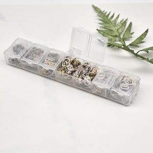 Op de afbeelding: Een doorzichtige plastic organizer gevuld met verschillende maten en kleuren van jumpringen. De jumpringen zijn zilver, goud en brons. Een groen varenblad staat op de achtergrond.