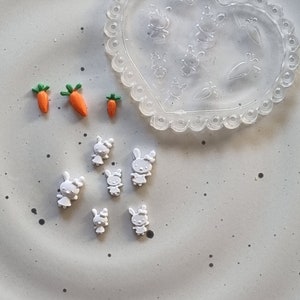 May include: A set of small white bunny and carrot shaped charms. The charms are arranged on a white surface. There is a clear plastic heart-shaped mould in the background.