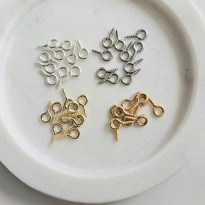 May include: A white plate with three piles of screw-eye hooks. The piles are separated into gold, silver, and nickel colours.