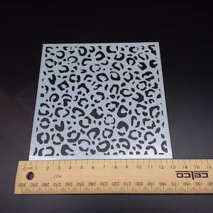 May include: A square stencil with a leopard print design. The stencil is made of a translucent material and features black spots. A wooden ruler with centimetre markings is placed next to the stencil.