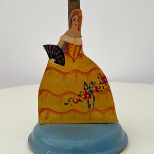 May include: A vintage paper doll toy with a painted woman in a yellow dress holding a black fan. The doll is attached to a blue metal base.