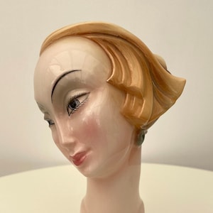 May include: A ceramic bust of a woman with a stylized blonde hairstyle. The bust is painted in shades of pink, white, and brown. The woman is wearing a green earring.