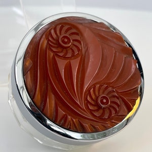 May include: A vintage, round, red-brown, plastic compact with a silver rim and a carved swirl design.