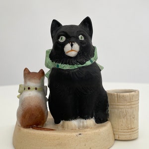 Peut inclure: Une figurine en céramique d'un chat noir aux yeux verts et un nœud vert, assis à côté d'un chat brun plus petit avec un nœud blanc. Les chats sont sur une base beige avec un petit tonneau en céramique blanc à droite.