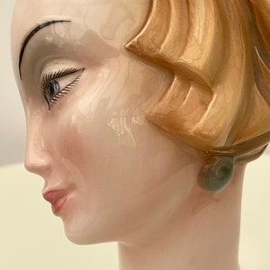 May include: A ceramic bust of a woman with a brown hairdo and a green earring. The bust is painted with realistic details, including a light pink blush on the cheeks and a light brown eyebrow.