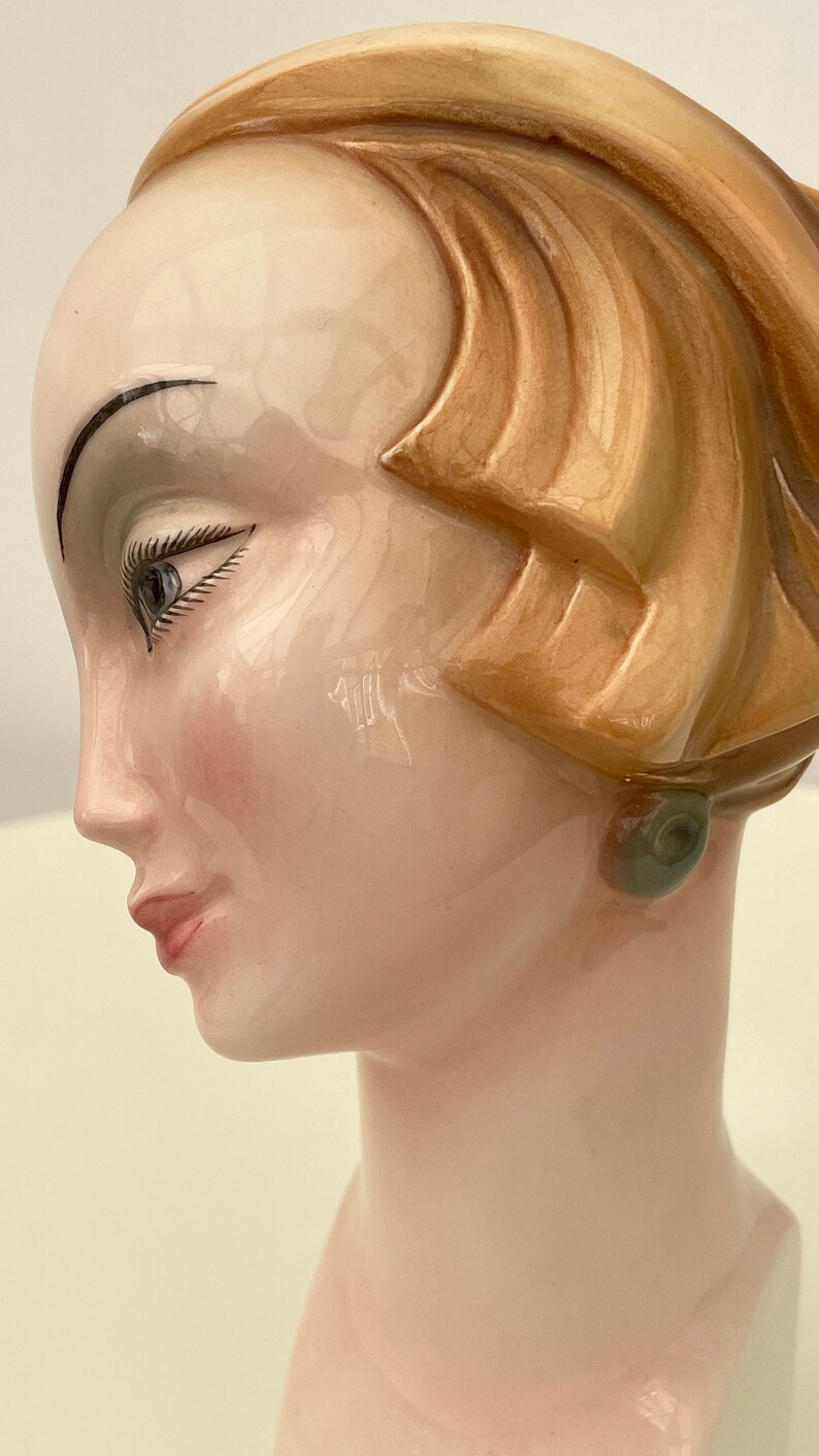 May include: A ceramic bust of a woman with a brown hairdo and a green earring. The bust is painted with realistic details, including a light pink blush on the cheeks and a light brown eyebrow.