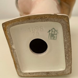 May include: White porcelain figurine of a woman with a brown hat. The figurine has a hole in the bottom and the base is marked with the number "1698 73" and a green house logo.