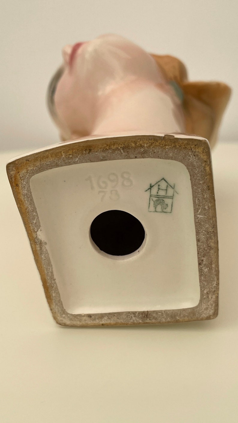 May include: White porcelain figurine of a woman with a brown hat. The figurine has a hole in the bottom and the base is marked with the number "1698 73" and a green house logo.