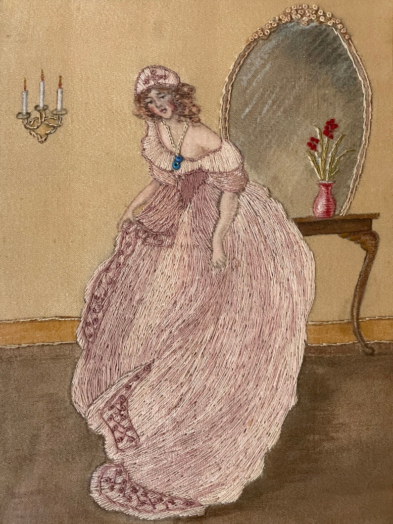 Puede incluir: Una imagen bordada de una mujer con un vestido rosa de pie frente a un espejo. Lleva un sombrero blanco y un collar con una piedra azul. Hay tres velas en un aplique de pared detr&aacute;s de ella.