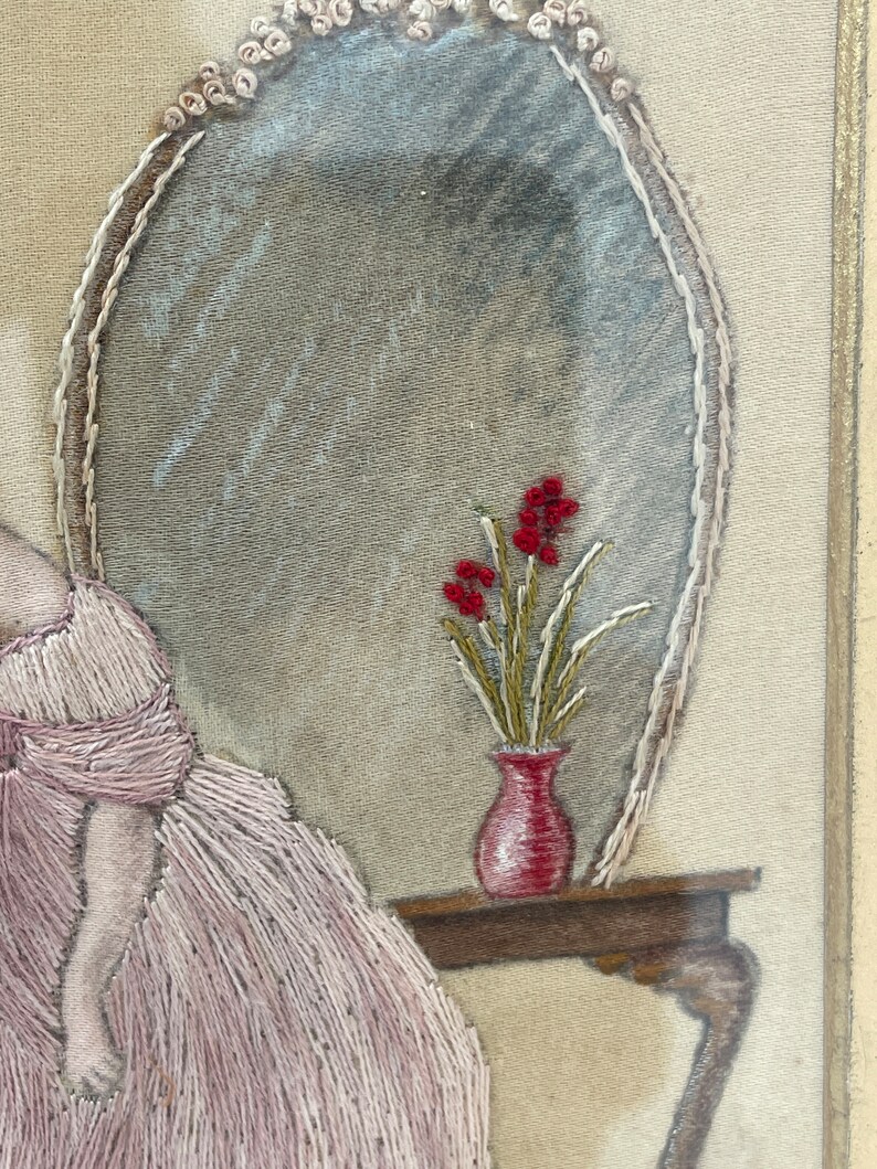 Puede incluir: Obra de arte bordada que representa a una mujer con un vestido rosa sentada junto a una mesa con un jarr&oacute;n de flores rojas. La obra est&aacute; enmarcada y tiene un aspecto vintage.