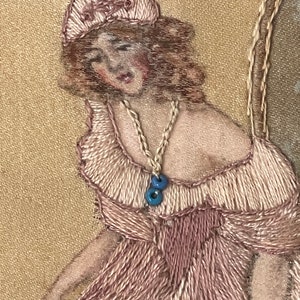 Puede incluir: Bordado que representa a una mujer con un vestido rosa y un sombrero blanco. Lleva un collar con cuentas azules. La obra est&aacute; realizada sobre una tela beige.