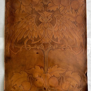 May include: A rectangular piece of tan leather featuring an embossed design of an owl perched on a branch. The owl is centered with wings spread wide, surrounded by detailed foliage and circular fruit motifs at the top and bottom edges.