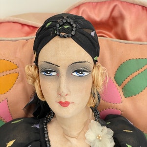 May include: A vintage cloth doll with blonde hair, wearing a black dress with a white flower accent, a black beaded necklace, and a black turban with a black beaded embellishment.