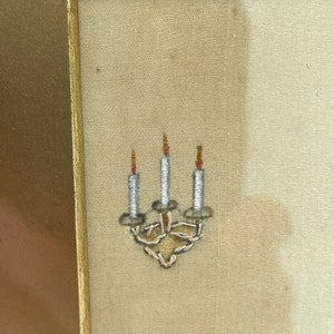 Puede incluir: Primer plano de un bordado enmarcado que representa un candelabro con tres velas. Las velas son blancas con llamas rojas y el candelabro est&aacute; hecho de hilo de plata.