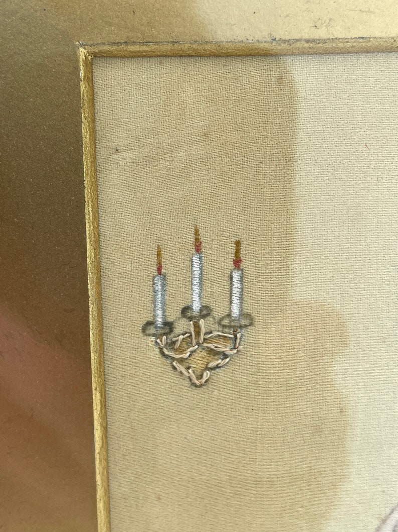 Puede incluir: Primer plano de un bordado enmarcado que representa un candelabro con tres velas. Las velas son blancas con llamas rojas y el candelabro est&aacute; hecho de hilo de plata.