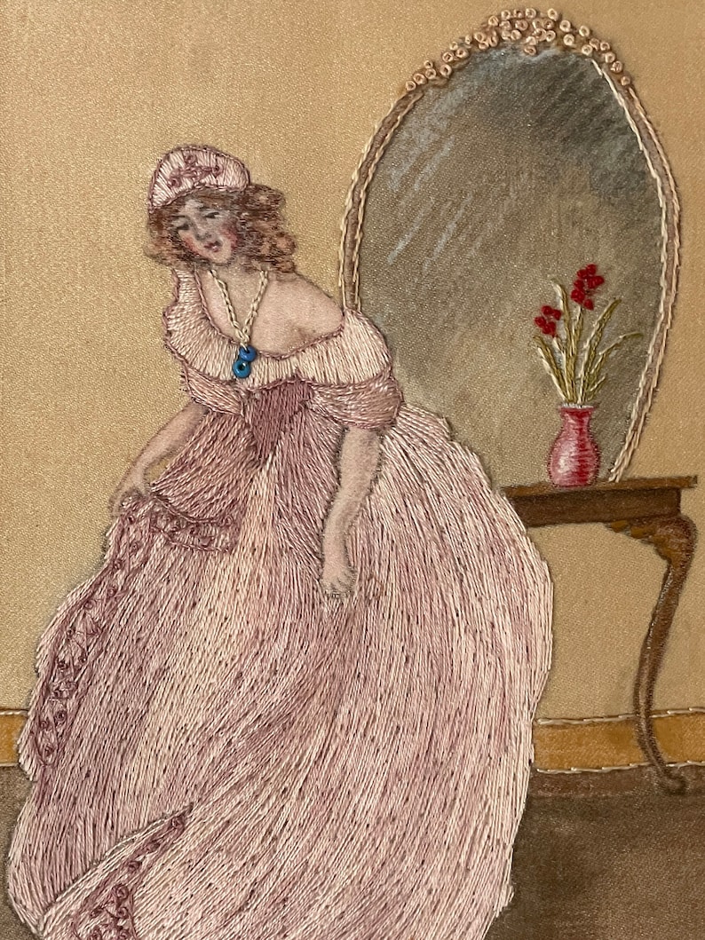 Puede incluir: Obra bordada que representa a una mujer con un vestido rosa de pie frente a un tocador con un espejo y un jarr&oacute;n de flores rojas. La mujer lleva una gorra blanca y un collar con un colgante azul.