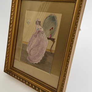 Puede incluir: Una obra de arte bordada enmarcada que representa a una mujer con un vestido rosa de pie frente a un espejo. La mujer sostiene una joya azul en su mano. La obra de arte est&aacute; enmarcada en un marco dorado con un borde decorativo.
