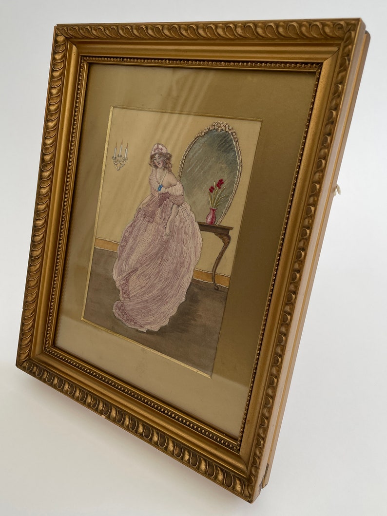 Puede incluir: Una obra de arte bordada enmarcada que representa a una mujer con un vestido rosa de pie frente a un espejo. La mujer sostiene una joya azul en su mano. La obra de arte est&aacute; enmarcada en un marco dorado con un borde decorativo.