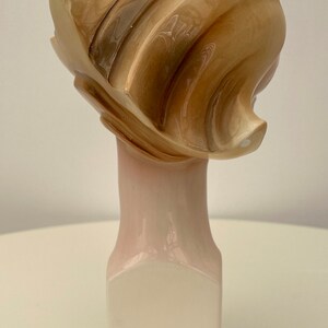 May include: A ceramic sculpture of a woman's head with a stylized, abstract hairstyle. The sculpture is painted in shades of brown and white and is mounted on a white pedestal.