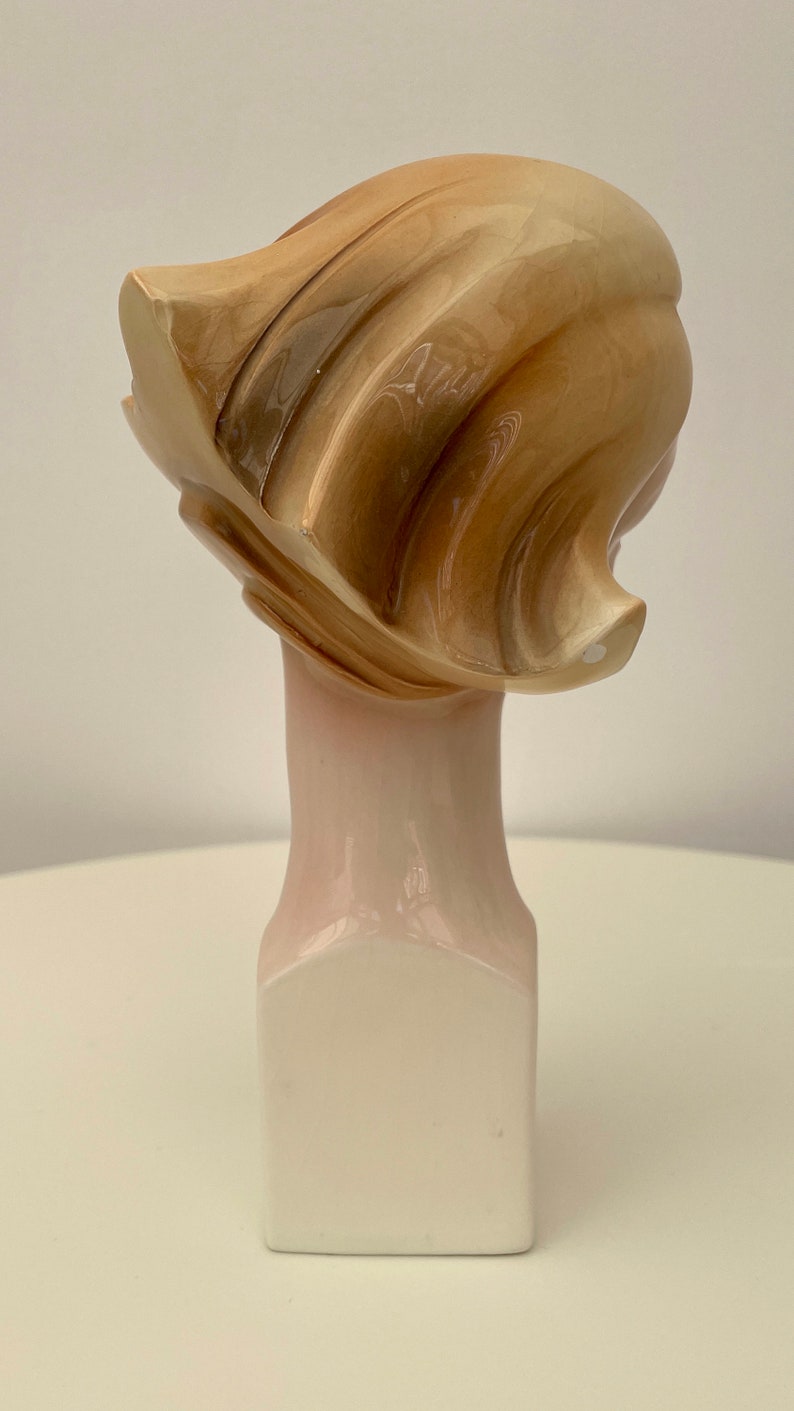 May include: A ceramic sculpture of a woman's head with a stylized, abstract hairstyle. The sculpture is painted in shades of brown and white and is mounted on a white pedestal.