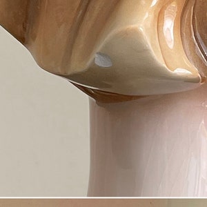 May include: A porcelain figurine of a woman with a brown and white dress. The figurine is in a standing position and has a slight smile on her face. The figurine has a small chip on the bottom.