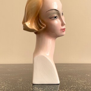 May include: A ceramic bust of a woman with blonde hair, wearing a white dress. The bust is painted in shades of pink and white.