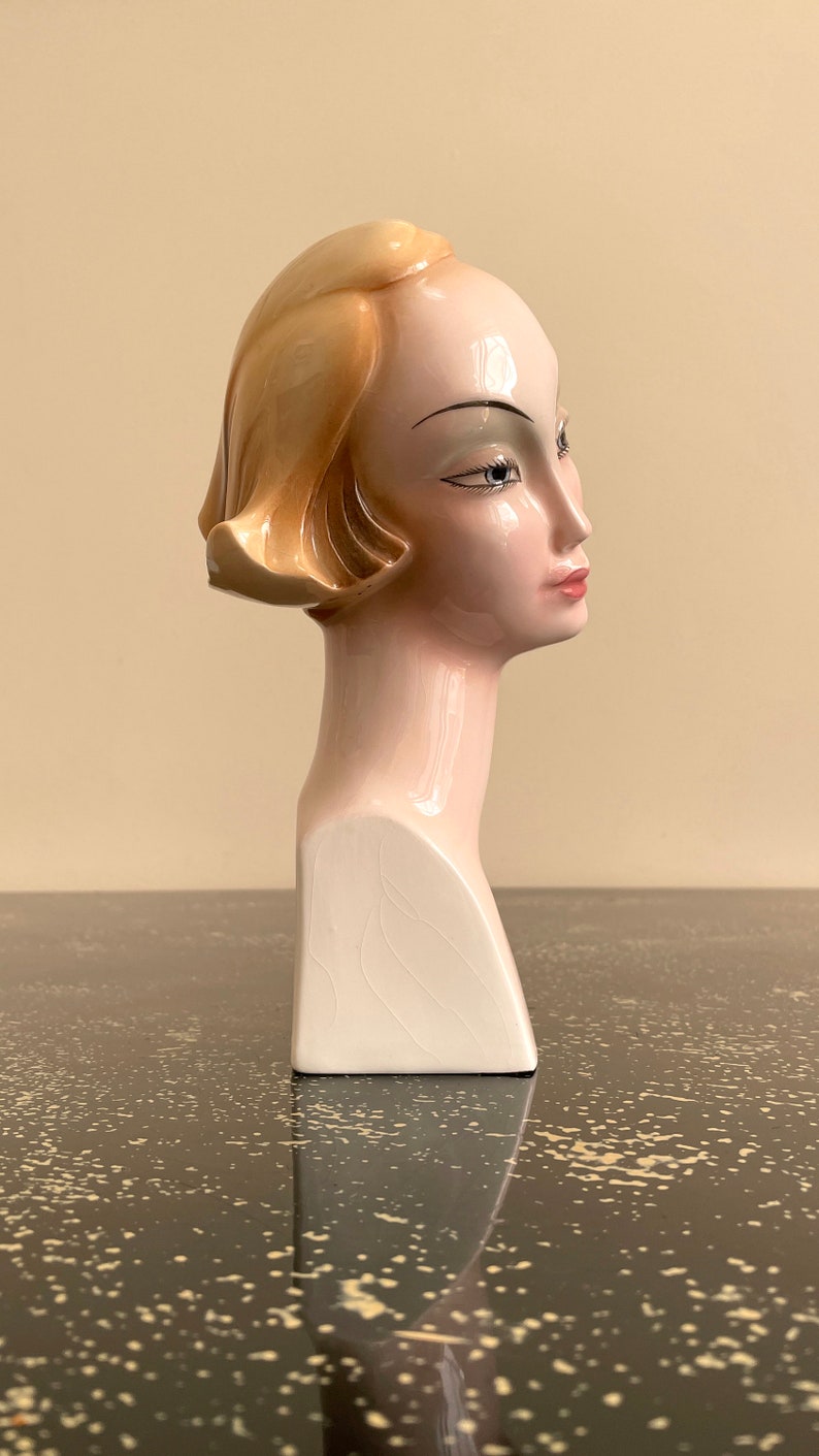 May include: A ceramic bust of a woman with blonde hair, wearing a white dress. The bust is painted in shades of pink and white.
