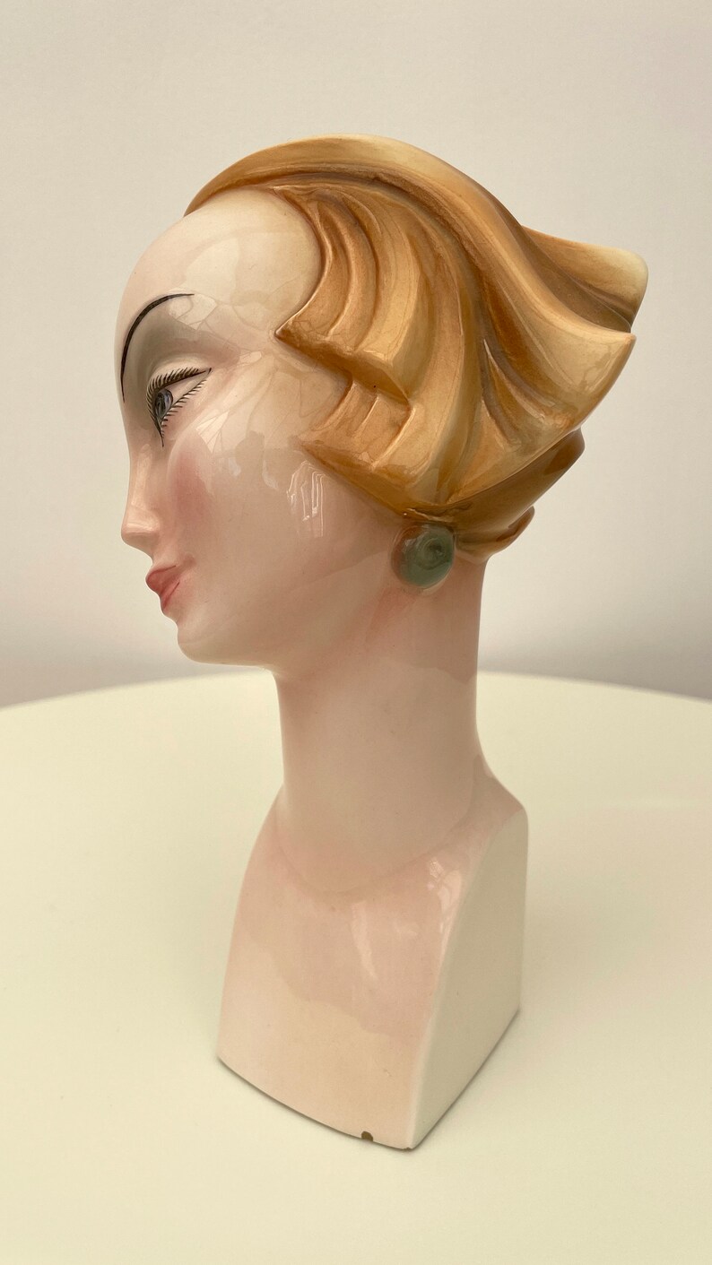 May include: A ceramic bust of a woman with a stylized hairstyle and a green earring. The bust is painted in shades of white, pink, and brown.